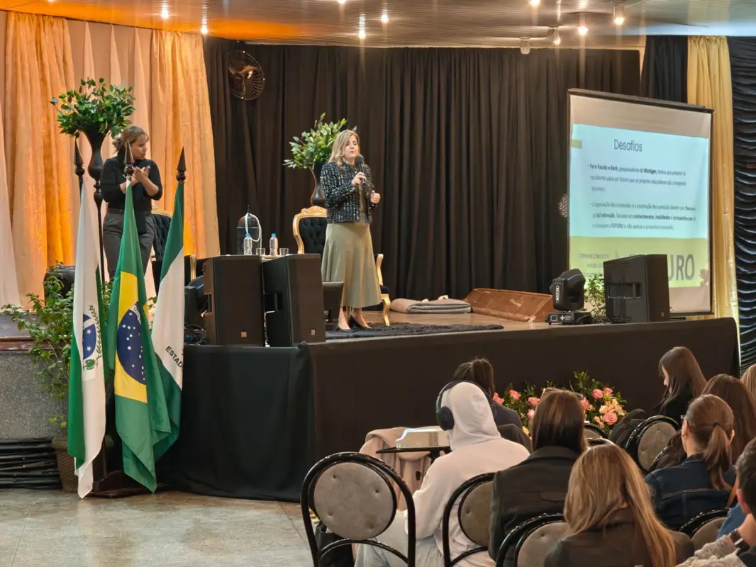 Marcia Baldini no palco de um salão, diante de educadores, com as bandeiras do Paraná e do Brasil ao fundo