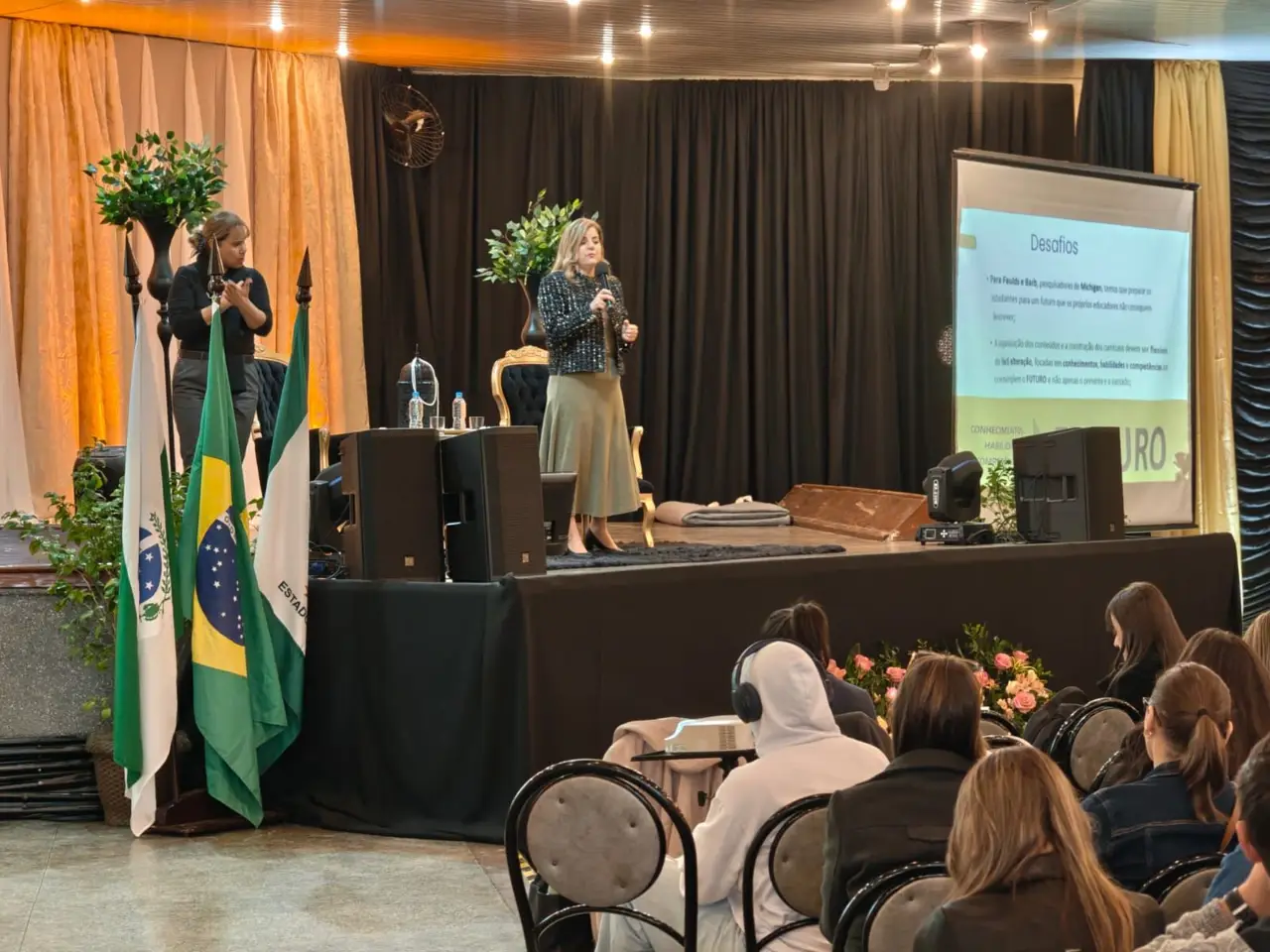Marcia Baldini no palco diante de educadores, com as bandeiras do Brasil e do Paraná