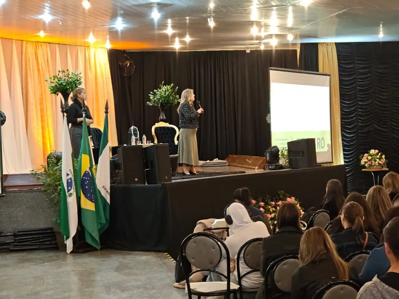 Marcia Baldini no palco, ao microfone, com as bandeiras do Paraná e do Brasil ao lado