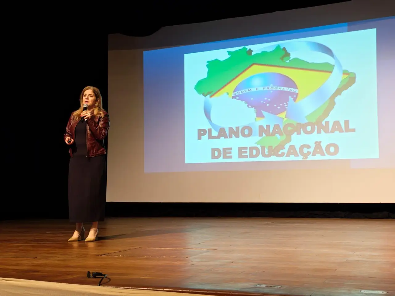 Marcia Baldini apresentando o Plano Nacional de Educação diante de um mapa do Brasil projetado