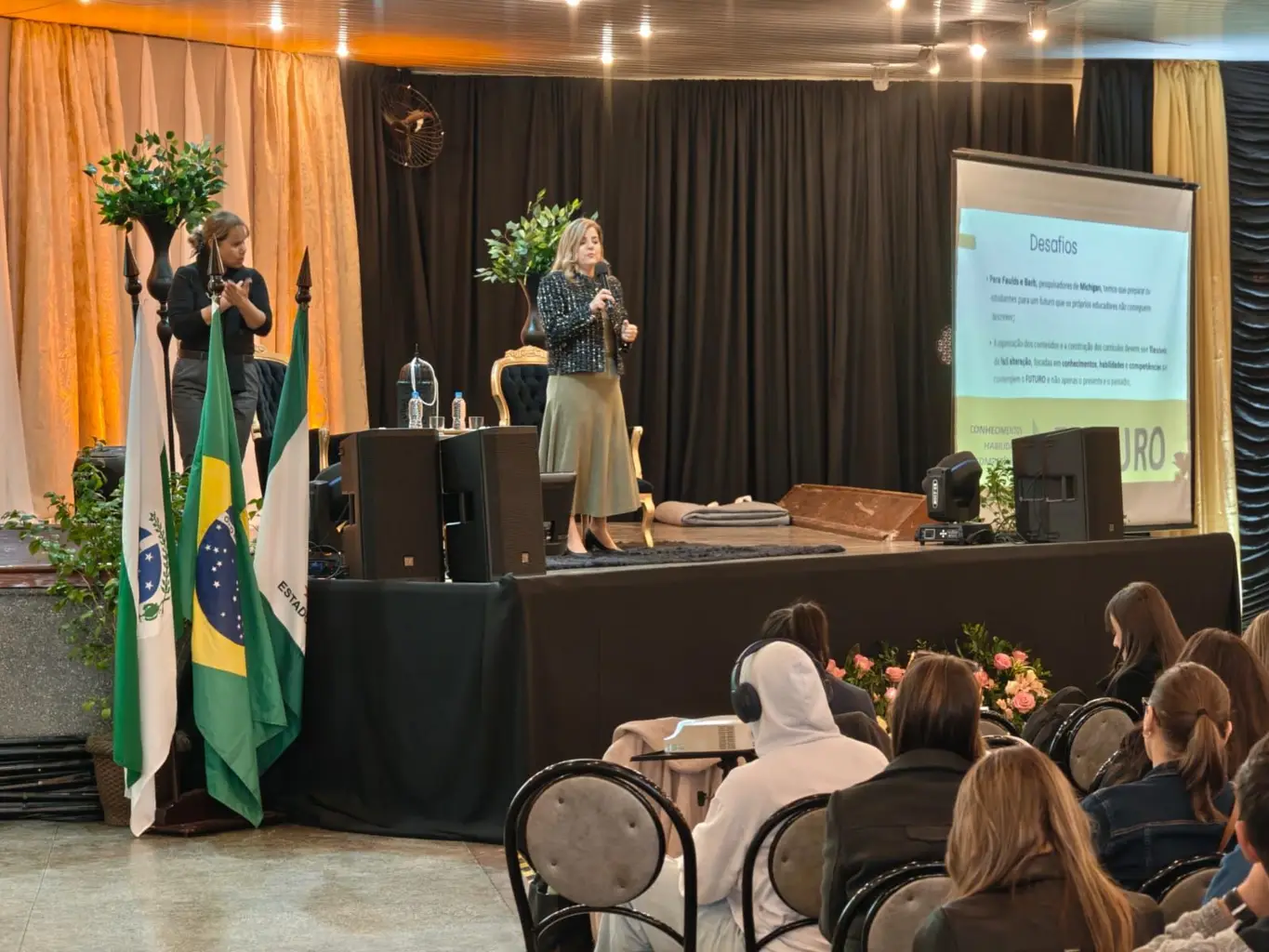 Marcia Baldini no palco de um salão, diante de educadores, com as bandeiras do Paraná e do Brasil ao fundo